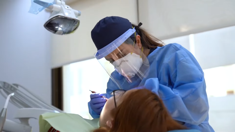 A Dentist Wearing Protective Gear Is Examining a Patient's Teeth Under a Bright Light, Highlighting Considerations Related to Dental Care Costs and Insurance Coverage